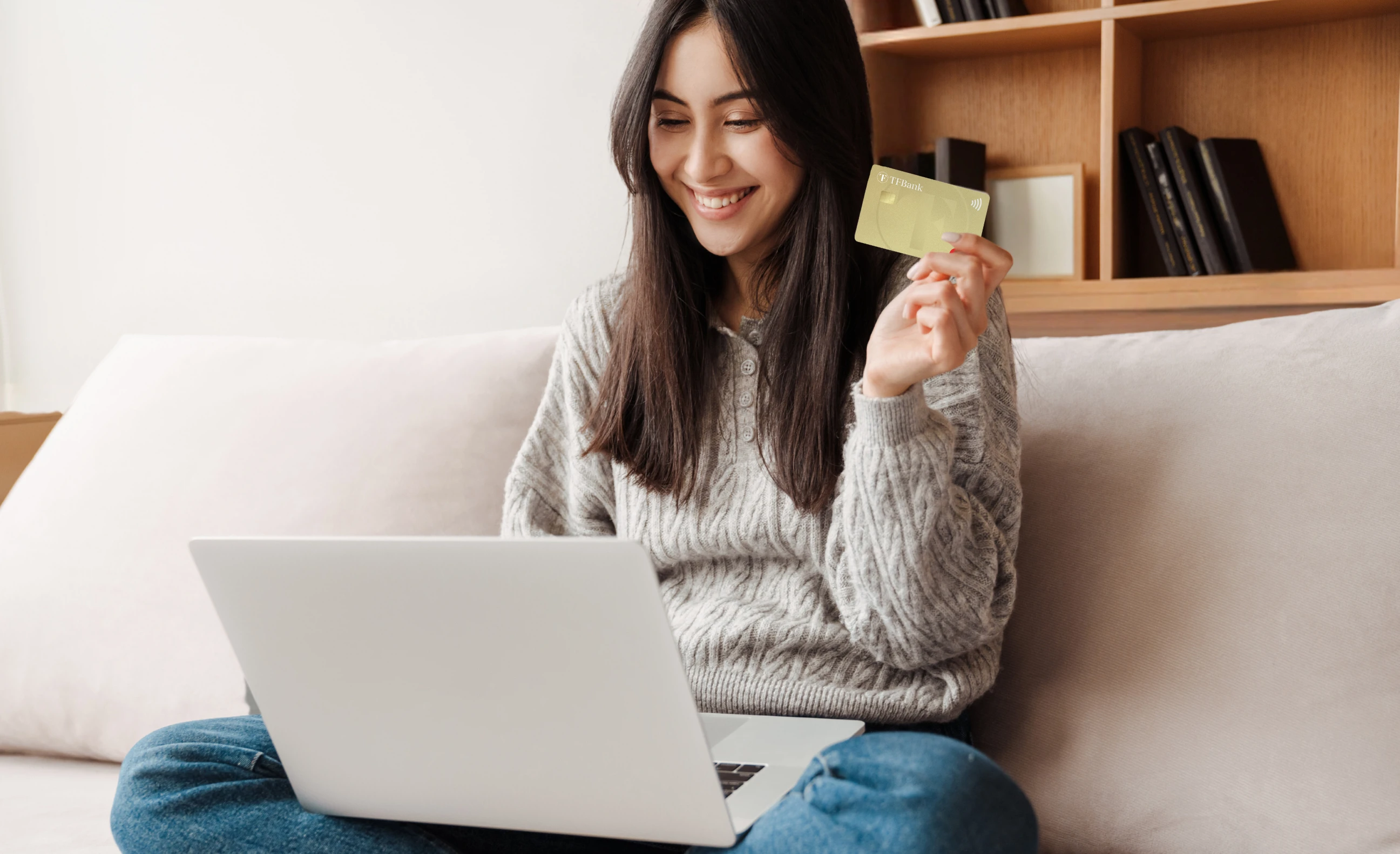 smilende kvinne sitter i sofa med pc i fanget og et tf bank mastercard i hånden