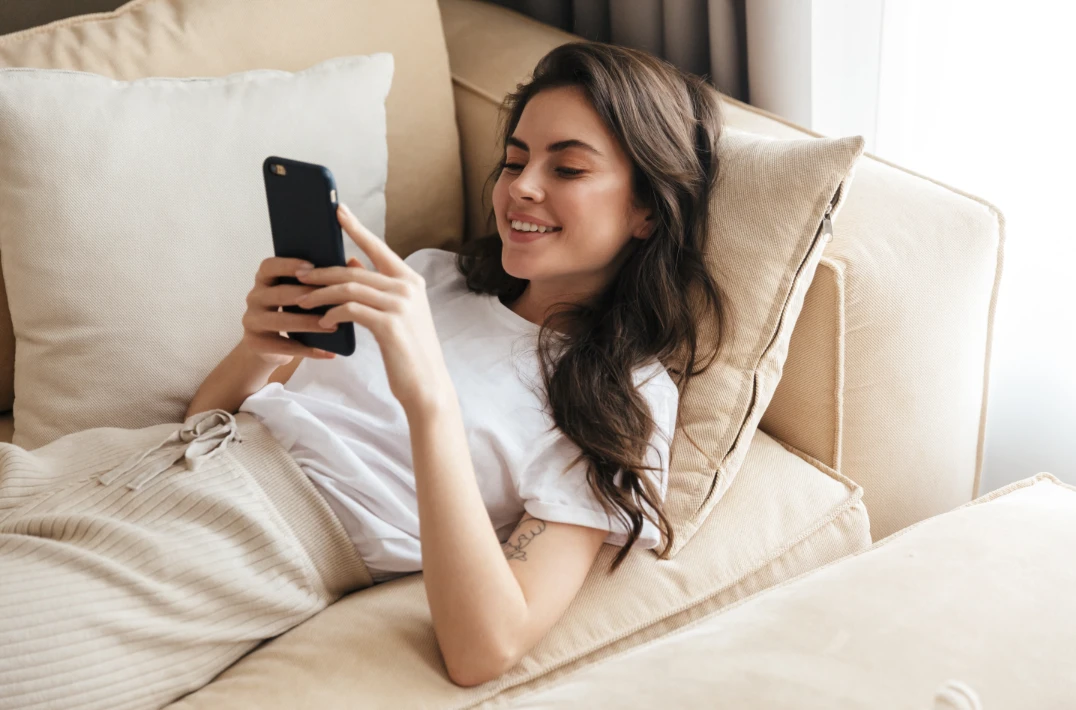 smilende kvinne ligger i sofa og ser på telefon